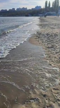 Güneşli bir sonbahar gününde insanlar sahilde yürüyor. Deniz kıyısının kumlu sahillerinde, kayan dalgaların yanında yürüyen insanlar yamaçta ağaçlar ve sonbahar günü binalar. Sonbaharda şehirde deniz. Dikey