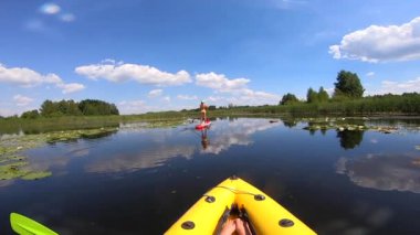 Güneşli bir yaz gününde, SUP tahtasının üzerinde yüzen bir kadının arkasında şişme bir teknede yüzen kişi. Sakin su bulutlu mavi gökyüzünü yansıtıyor. Doğa. Spor Gevşetici Turizm Seyahat arka planı