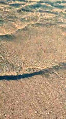 Güneşli bir günde denizin kumsalındaki temiz şeffaf su dalgalarının soyut görüntüsü. Gölgeli dalgalar kumsala yakın çekim yapıyor. Kumlu zeminde güneş parlıyor. Dikey