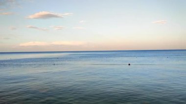Akşam denizi sakin görünüyor. Pembe turuncu gökyüzü ve akşam vakti beyaz bulutlar. Deniz suyunun pürüzsüz mavi yüzeyi. Suda kısıtlı şamandıralar var. Sakin bir hava. Soyut doğal arkaplan