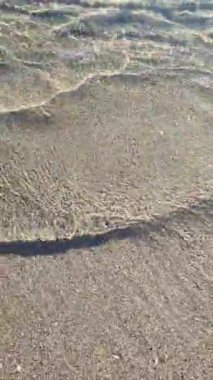 Kumsalda kumsalda yuvarlanan küçük deniz dalgalarının temiz şeffaf sularının soyut görüntüsü. Deniz dalgalarının gölgeleri ve kumlu zeminde su yüzeyinden parlayan güneş. Doğal arkaplan dikey