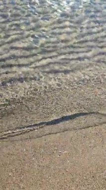 Güneşli bir günde, küçük şeffaf su dalgaları sahilin kumlarına yuvarlanıyor. Güneş parlıyor, su yüzeyinin deniz tabanındaki yansıması. Gölgelerin soyut çizgileri kumun üzerindeki su. En alttaki tepeler Dikey