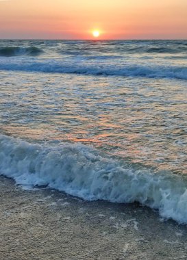 Fırtınalı deniz. Gün batımında ve şafakta denizde beyaz köpüklü büyük dalgalar. Mavi deniz, turuncu kırmızı gökyüzü ve güneş. Sahil kıyısı kıyı şeridi. Doğal arka plan doğa fırtınası