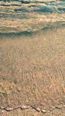 Kumsalda kumsalda yuvarlanan küçük deniz dalgalarının temiz şeffaf sularının soyut görüntüsü. Deniz dalgalarının gölgeleri ve kumlu zeminde su yüzeyinden parlayan güneş. Doğal arkaplan dikey