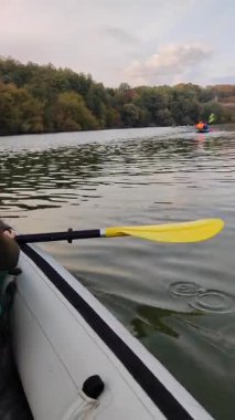 Şişme kayıkta oturan kadın kayık gri renkte, suda sarı kürek ve sonbahar günü büyüyen ağaçla kıyıya yakın yüzüyor. Arka planda yüzen bir sürü kano teknesi var. Dikey