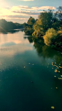 Doğa manzarası. Mavi nehir, kıyılardaki ağaçlar, mavi gökyüzü, beyaz bulutlar ve parlayan güneş. Rüzgâr ve dalgalar esiyor suyun yüzeyinde..