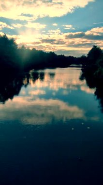 Nehir ve ağaçların yeşil yaprakları kıyılarda, gökyüzünde ve gün batımında ve şafakta beyaz bulutlarda büyüyor. Sakin, rüzgarsız hava. Doğa. Doğal arka plan. Çevre