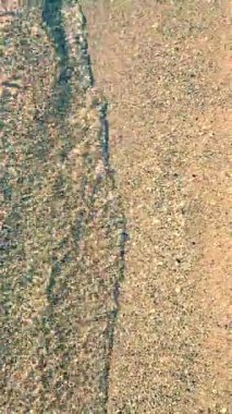 Kumsalda kumsalda yuvarlanan küçük deniz dalgalarının temiz şeffaf sularının soyut görüntüsü. Deniz dalgalarının gölgeleri ve kumlu zeminde su yüzeyinden parlayan güneş. Doğal arkaplan dikey