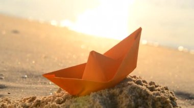 Paper orange boat on the sand near the sea waves with sunny path on sandy beach of sea shore during sunset dawn with shining sun close-up. Swimming dream sailing traveling vacation tourism concept