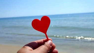 Parmaklarını tutan kişi arka planda kırmızı kalp, deniz ve deniz dalgaları, güneşli yaz günlerinde deniz kenarında mavi gökyüzü. Aşk romantizmini Aziz Sevgililer Günü olarak kabul et.