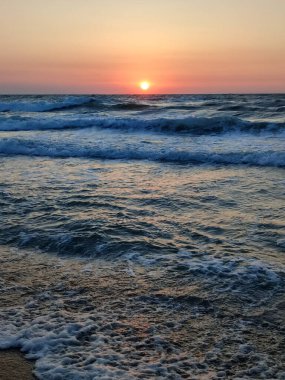 Fırtınalı deniz. Gün batımında ve şafakta denizde beyaz köpüklü büyük dalgalar. Mavi deniz, turuncu kırmızı gökyüzü ve güneş. Sahil kıyısı kıyı şeridi. Doğal arka plan doğa fırtınası