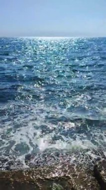 Güneşli sıcak bir günde güzel, temiz bir deniz. Mavi sakin deniz yüzeyinde güneş parlıyor ve mavi gökyüzü. Beyaz köpüklü deniz dalgaları kıyıdaki eski taşlara çarpıyor. Doğa deniz manzarası, deniz manzarası. Dikey