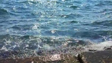 Güneşli sıcak bir günde güzel, temiz bir deniz. Mavi sakin deniz yüzeyinde güneş parlıyor ve mavi gökyüzü. Beyaz köpüklü deniz dalgaları kıyıdaki eski taşlara vuruyor ve çarpıyor. Doğa deniz manzarası, deniz manzarası..