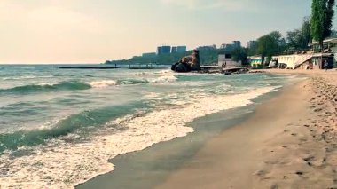 Deniz kıyısının kumlu sahillerinde yürüyen insanların ve güneşli bir günde deniz kıyısında binaların olduğu güzel bir deniz manzarası. Beyaz köpüklü deniz dalgaları, güneşli bir günde kumlu bir kıyıda büyük bir kaya ve insanlarla yuvarlanıyor.