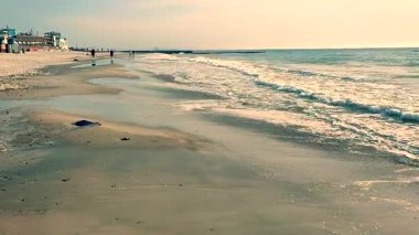 Deniz kıyısının kumlu sahillerinde yürüyen insanların ve güneşli bir günde deniz kıyısında binaların olduğu güzel bir deniz manzarası. Beyaz köpüklü deniz dalgaları güneşli bir günde insanlarla kumlu bir kıyıya vuruyor.