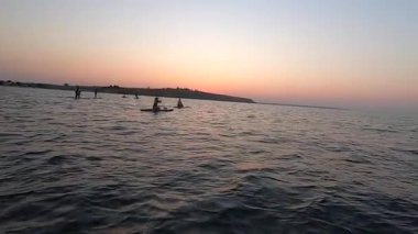 Şafak vakti SUP sörf tahtasında yüzen bir adam. Beyaz SUP tahtasının üzerinde yüzen ve günbatımında kıyıya yakın dalgalarla kürek çeken insanlar. Aktif yaşam tarzı eğlence sporu POV