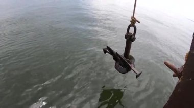 Denizde giden bir geminin gövdesinde sallanan eski bir çapa. Eski paslı çapa ipte sallanıyor ve denizin üstünde yüzen ahşap bir geminin pruvasının yanında sendeliyor. Seyahat turizmi