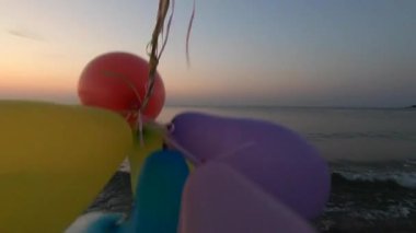 Alacakaranlıkta deniz kıyısında renkli balonlarla oynayan biri. Gün batımında deniz sahilinde bir sürü renkli balon elinde. Çok renkli balonlar kurdeleyle bağlanmış. Yavaş çekim
