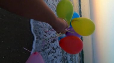 Alacakaranlıkta deniz kıyısında renkli balonlarla oynayan biri. Gün batımında deniz sahilinde bir sürü renkli balon elinde. Çok renkli balonlar kurdele bağladı. Ağır çekim. Dikey