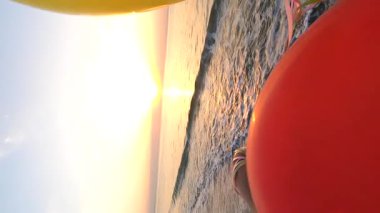 Deniz kıyısında renkli balonlarla oynayan bir adam var. Gün batımında deniz sahilinde bir sürü renkli balon elinde. Güneş topların arasından parlıyor. Ağır çekim. Dikey