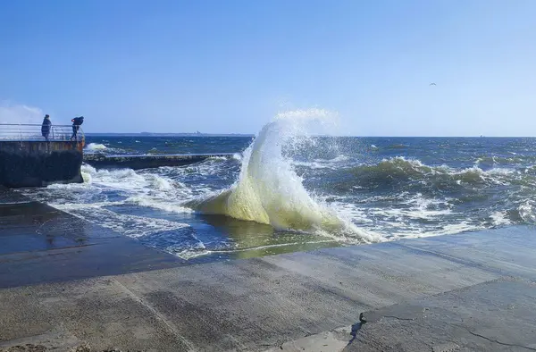Güneşli bir yaz gününde denizde fırtına. Güneşli bir günde beton eski iskeleye beyaz köpüklü büyük deniz dalgaları çarpıyor. Çimento iskelesine ve temiz mavi gökyüzüne çarpan büyük dalgalardan bir sürü sıçrama.