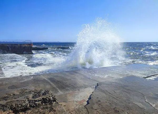 Güneşli bir yaz gününde denizde fırtına. Güneşli bir günde beton eski iskeleye beyaz köpüklü büyük deniz dalgaları çarpıyor. Çimento iskelesine ve temiz mavi gökyüzüne çarpan büyük dalgalardan bir sürü sıçrama.