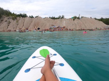 Taş ocağı gölünün mavi sularında yüzen SUP tahtasıyla kayalık kıyılar ve yaz günü kürek çeken ağaçlar. Yaz sporları, turizm, dinlenme, rahatlama, seyahat, dinlenme