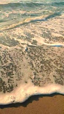 Beyaz köpüklü deniz dalgaları kumlu sahil kıyısında yuvarlanıyor. Deniz kıyısındaki sarı kumdaki açık mavi deniz dalgalarının görüntüsü. Deniz Burnu. Deniz manzarası. Kıyı manzarası Doğa Doğal arka plan Dikey