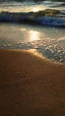 Güneş doğarken ya da batarken kumların üzerinde yuvarlanan güzel deniz dalgaları. Beyaz köpüklü mavi deniz suyunun sakin dalgaları ve güneşli bir patika ve ıslak kumun üzerine sıçrıyor. Tatil turizmi. Dikey