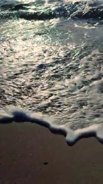 Güneş doğarken ya da batarken kumların üzerinde yuvarlanan güzel deniz dalgaları. Beyaz köpüklü mavi deniz suyunun sakin dalgaları ve güneşli bir patika ve ıslak kumun üzerine sıçrıyor. Tatil turizmi. Dikey