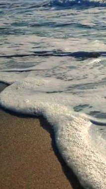 Deniz kıyısındaki kumların üzerinde dalgalar yuvarlanıyor. Beyaz köpüklü mavi deniz suyunun sakin dalgaları ve ıslak kumun üzerine su sıçratır. Tatil turizmi. Dikey