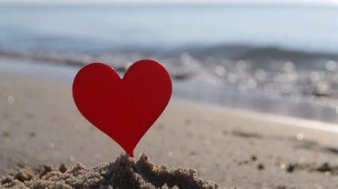 Plastik çubuk şeklinde kırmızı kalp, kumlu sahilde, deniz dalgalarının arka planında güneşli yaz günlerinde. Aşk ilişkisini kavramsallaştır. Aşk aşkı. Sevgililer Günü 'nü sev.