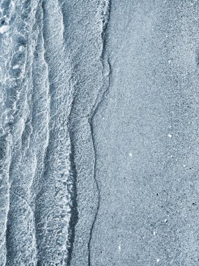 Güneşli bir günde, deniz kıyısının kumlu sahilinde güzel, şeffaf, küçük deniz dalgaları. Temiz su, güneş parlaması ve yakın plan kumlu zeminde gölgeler. Doğal soyut arkaplan.