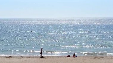 Güneşli bir yaz gününde insanlarla birlikte deniz ve kumsal manzarası. Panoramik mavi deniz, durgun ve sıcak havada, beyaz güneş, su yüzeyinde parlıyor. İnsanlar kumsalda güneşleniyor ve dinleniyor.