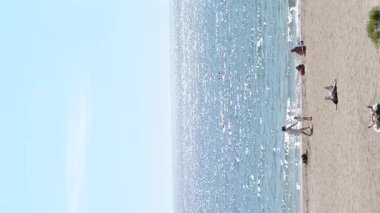 Deniz manzarası, mavi gökyüzü ve güneşli yaz gününde insanlarla dolu kumlu plaj. Panoramik mavi deniz, durgun ve sıcak havada, beyaz güneş, su yüzeyinde parlıyor. İnsanlar kumda güneşleniyor. Dikey