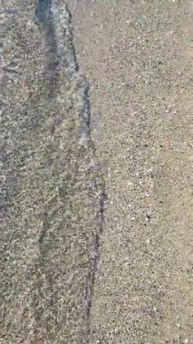 Güneşli bir günde, deniz kıyısının kumlu sahilinde güzel, şeffaf, küçük deniz dalgaları. Temiz su, güneş parlaması ve yakın plan kumlu zeminde gölgeler. Doğal soyut arka plan. Dikey