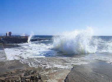Güneşli bir yaz gününde denizde fırtına. Güneşli bir günde beton eski iskeleye beyaz köpüklü büyük deniz dalgaları çarpıyor. Çimento iskelesine ve temiz mavi gökyüzüne çarpan büyük dalgalardan bir sürü sıçrama.
