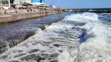 Odessa, Ukrayna, 30 Ağustos 2023: Beyaz köpüklü büyük dalga birçok insanın bulunduğu eski çimento kıyısına çarpıyor. Güneşli bir günde dalgalar iskeleye vuruyor. Fırtına, fırtınalı deniz.
