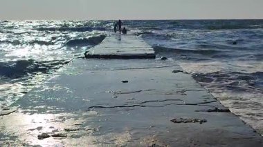 Güneşli bir günde fırtınalı bir denizde ıslak bir iskelede duran insanların siyah siluetleri. Dalgalar iskeleye çarpıyor. Bir sürü büyük sıçrama.
