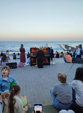 Odessa, Ukrayna, 25 Mayıs 2024: Piyanist deniz kenarında arka planda piyano çalıyor ve yaz gecesi onu dinleyen birçok kişi var. Seyircilerle piyano çalan müzisyen konseri.