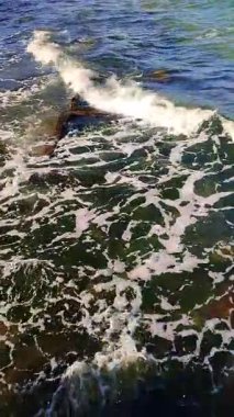 Beyaz köpüklü mavi deniz dalgaları kıyıda yuvarlanıyor. Suyun altında yeşil yosunlarla kaplanmış eski beton levhalar. Soyut deniz arka planı. Doğal arka plan. Doğa. Deniz kıyısında. Dikey