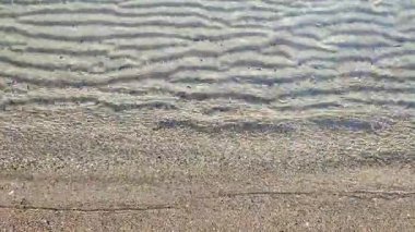 Güneşli bir günde, deniz kıyısının kumlu sahilinde, şeffaf, küçük deniz dalgaları. Temiz su, güneş parlaması ve yakın plan kumlu zeminde gölgeler. Doğal soyut arkaplan