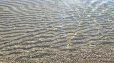 Güneşli bir günde, deniz kıyısının kumlu sahilinde, şeffaf, küçük deniz dalgaları. Temiz su, güneş parlaması ve yakın plan kumlu zeminde gölgeler. Doğal soyut arkaplan