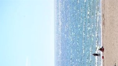 Deniz manzarası, mavi gökyüzü ve güneşli yaz gününde insanlarla dolu kumlu plaj. Panoramik mavi deniz, durgun ve sıcak havada, beyaz güneş, su yüzeyinde parlıyor. İnsanlar kumda güneşleniyor. Dikey