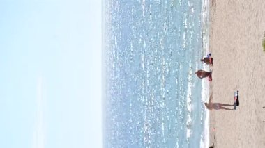 Deniz manzarası, mavi gökyüzü ve güneşli yaz gününde insanlarla dolu kumlu plaj. Panoramik mavi deniz, durgun ve sıcak havada, beyaz güneş, su yüzeyinde parlıyor. İnsanlar kumda güneşleniyor. Dikey