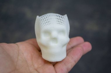 Elinde erimiş plastikten siyah zemin üzerine basılmış insan kafatasının küçük beyaz prototipini tutan kişi. Üç boyutlu model. Katkı teknolojisi. Modern yeni baskı endüstrisi.