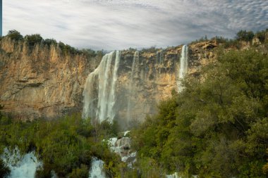 Scenic view of a waterfall flowing down a rocky cliff surrounded by a dense forest, creating a breathtaking natural landscape clipart