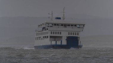 Dalgalı bir denizde yelken açan feribot. Arka planda bir rüzgar türbini çiftliği var.