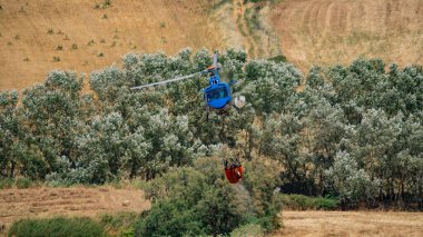 İtfaiye helikopteri, rüzgar türbini ve orman tepelerinin üzerinde su kovası ile uçar.