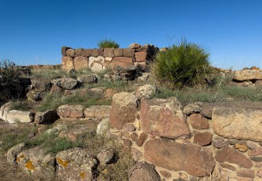Monte Sirai 'nin arkeolojik taş kalıntıları ve açık güneşli gökyüzünün altında vahşi bitki örtüsü.